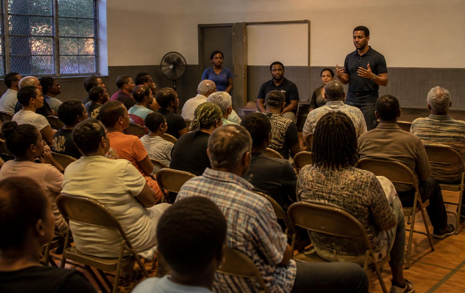 Residents of many ages seated in folding chairs listening to a speaker at a community meeting